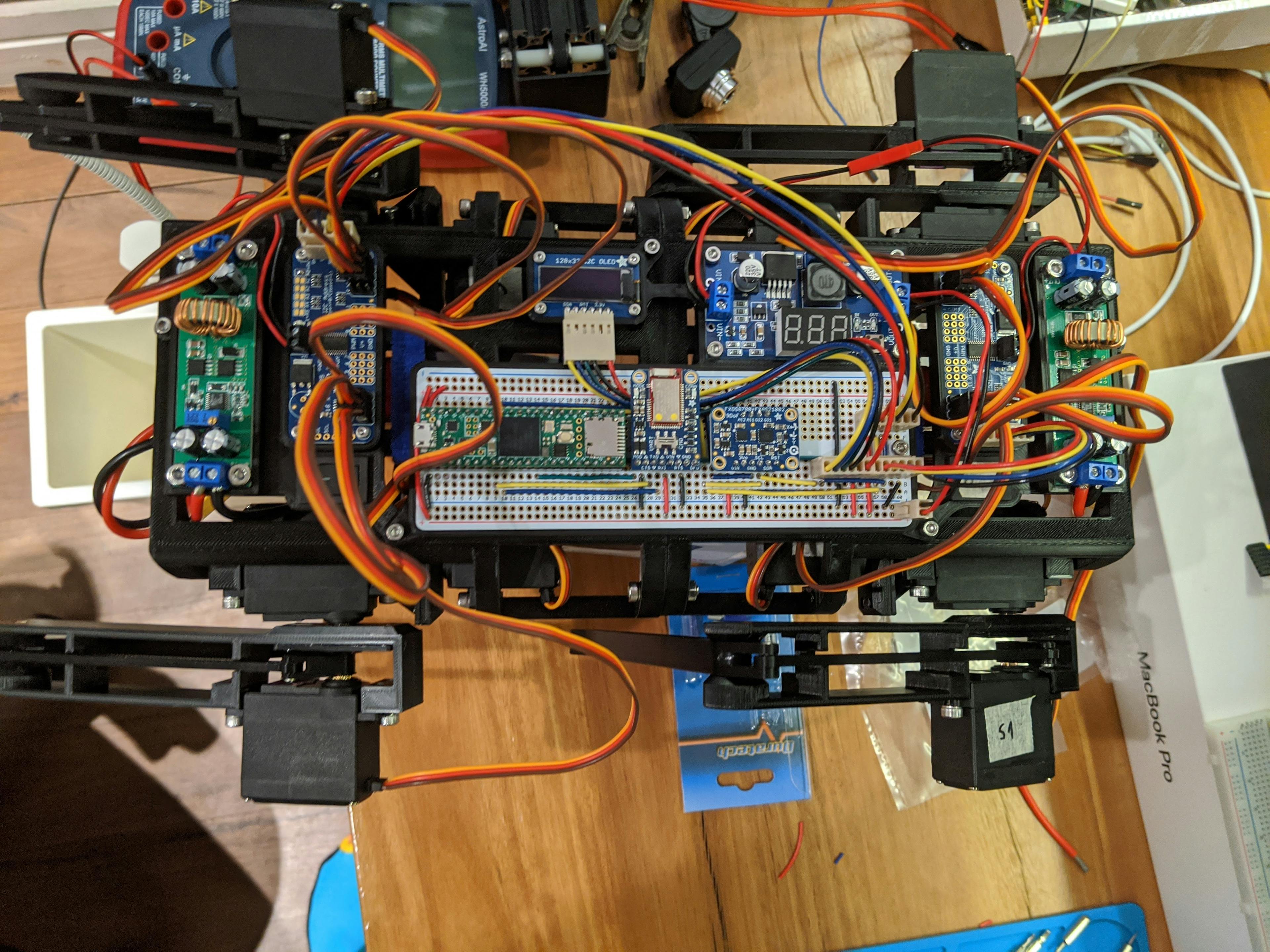 Top-down view of the quadruped. Cat-sized, 3D printed, black frame on which 9 PCBs are mounted interconnected with various colorful cables.